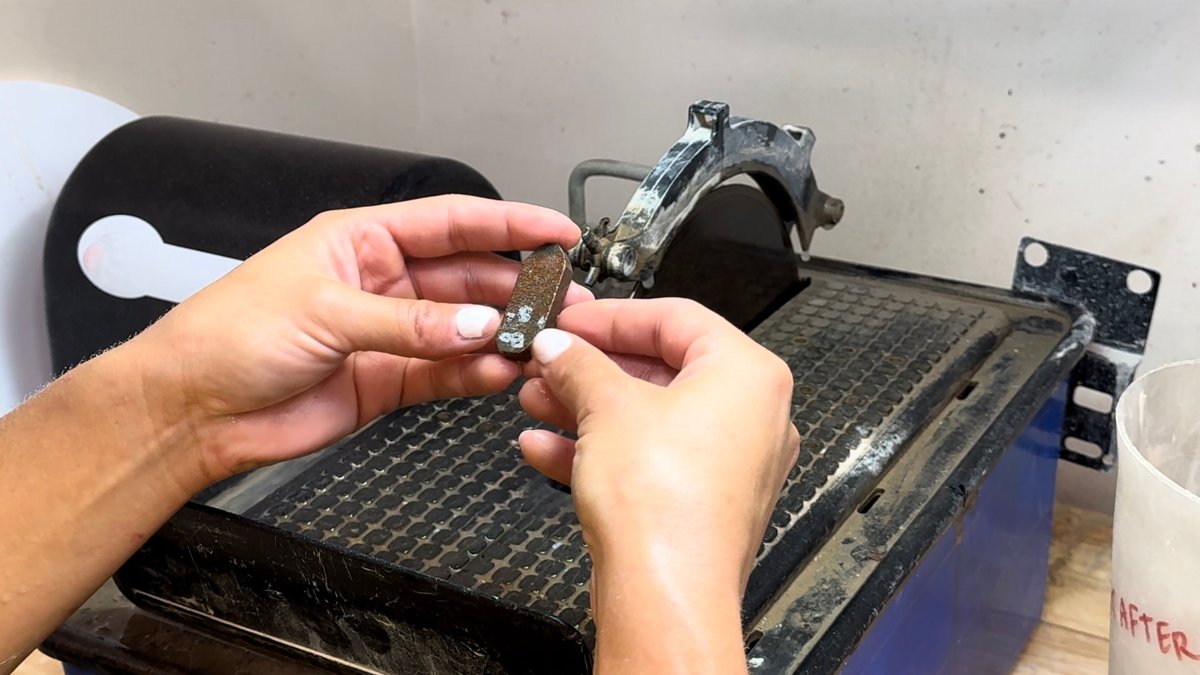 A student holds a small slab of stone up to the blade of a lapidary trim saw in the B Golden Jewelry School studio, from the official Intro to Lapidary class page.