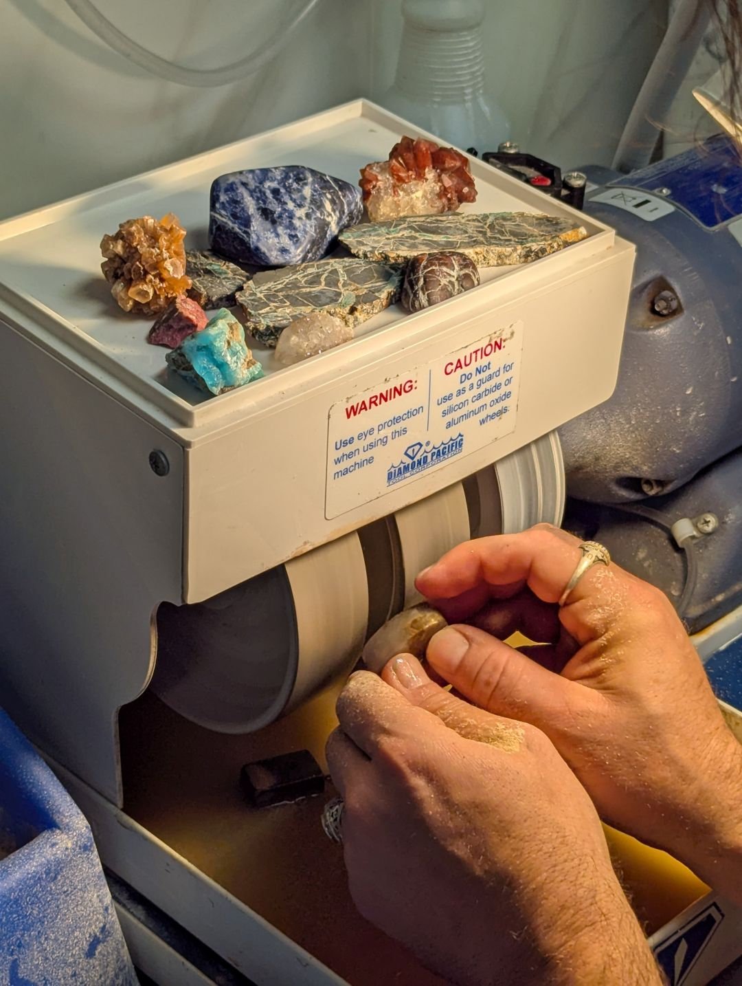 Hands shaping a stone on a Diamond Pacific cabbing machine in the DIYcave lapidary room, with rough and slabbed stones on the machine hood, from the official DIYcave class page.