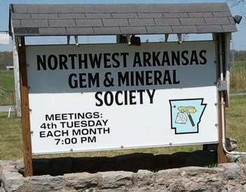 Outdoor clubhouse sign reading Northwest Arkansas Gem & Mineral Society, Meetings: 4th Tuesday each month 7:00 PM, with an Arkansas-shaped emblem showing a rock hammer, set on a stone base under a blue sky, from the official Northwest Arkansas Gem and Mineral Society website.