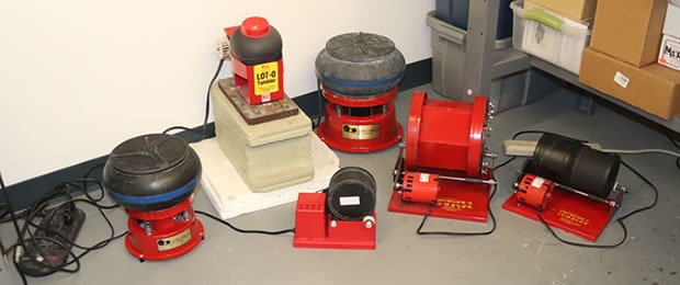 A row of red rock tumblers on a gray warehouse floor, including vibratory tumblers with black bowls, a Lot-O Tumbler on a stone block and Thumler's rotary tumblers with black rubber barrels, from the official RockTumbler.com About page.