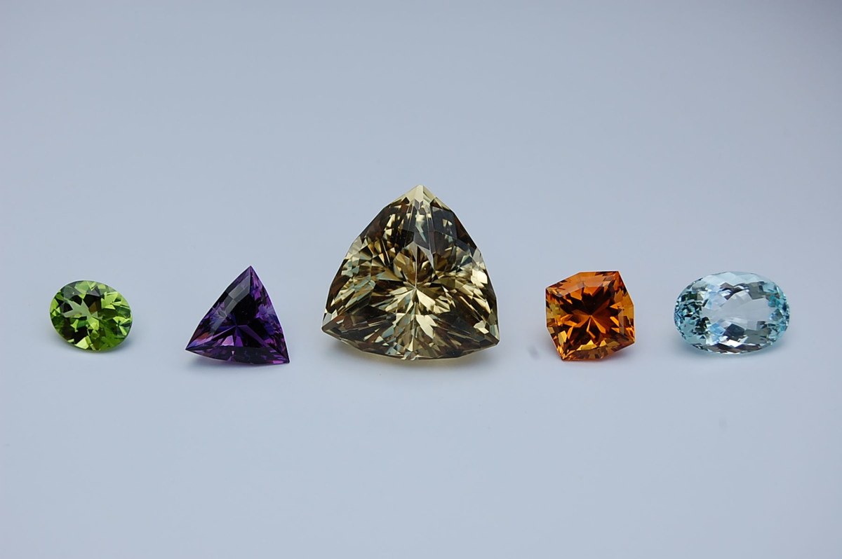 Five faceted gemstones in a row on a pale gray background: a green oval, a purple triangle, a large golden-brown triangle, an orange square and a pale blue oval, from the official St. Louis Mineral and Gem Society website.